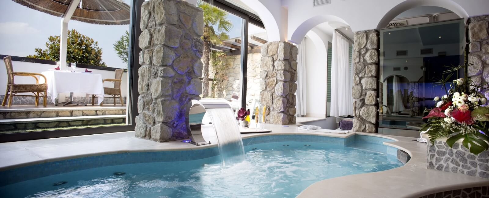 Motel K FantaSuite Antigua vista su piscina idromassaggio con cascata, sullo sfondo si intravede la terrazza esterna con tavolo ristorazione sotto al gazebo estivo, camera a tema isole dei caraibi